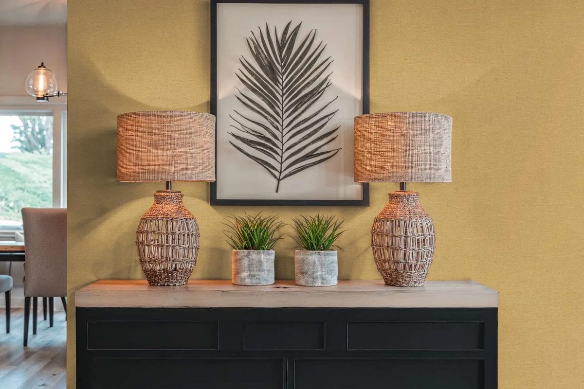 Dzeltenas spīdīgas neaustas tapetes ar dabisku tekstila dizainu saulainā guļamistabas interjerā. SKU: 3012157, AS Creation.